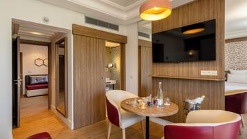 A modern hotel room with a wooden wall panel, a flat-screen TV, a round wooden table with a bottle of wine, and a red leather sofa.