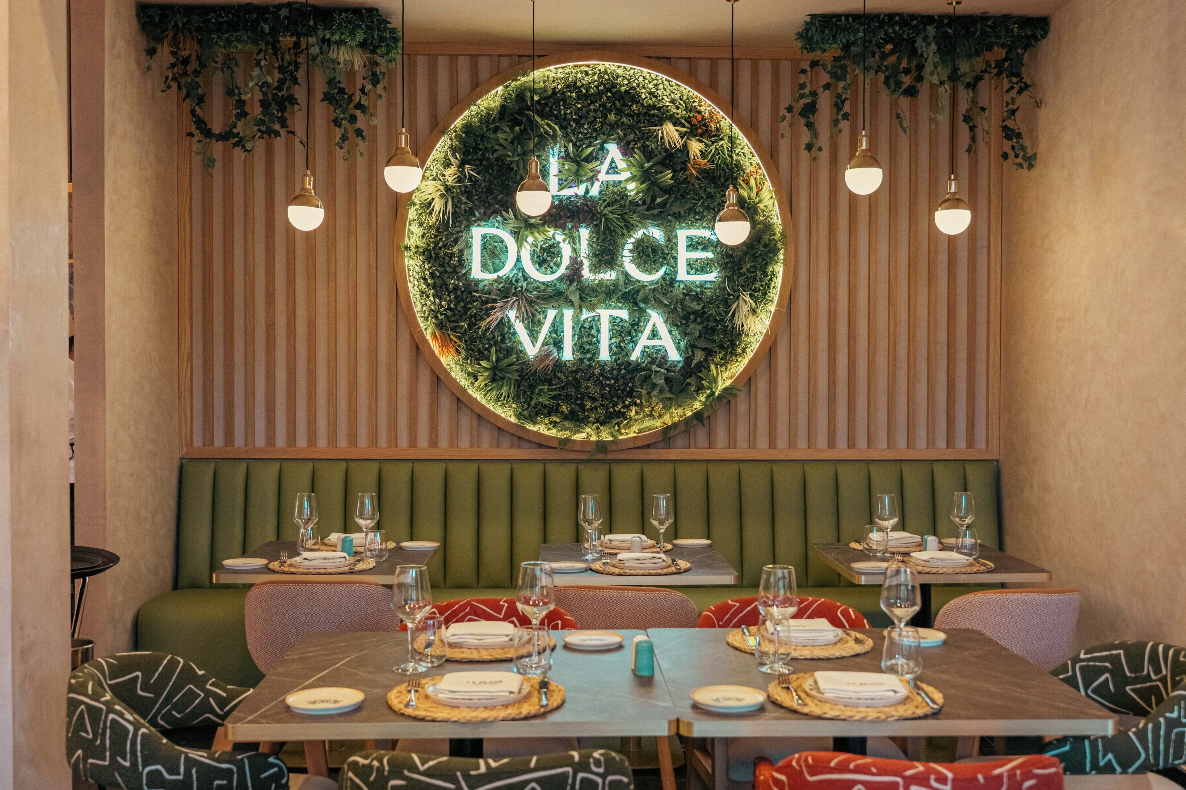 A restaurant interior with a neon sign reading 'LA DOLCE VITA,' green upholstered booths, and tables set with plates and glasses.