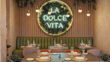 A restaurant interior with a neon sign reading 'LA DOLCE VITA,' green upholstered booths, and tables set with plates and glasses.
