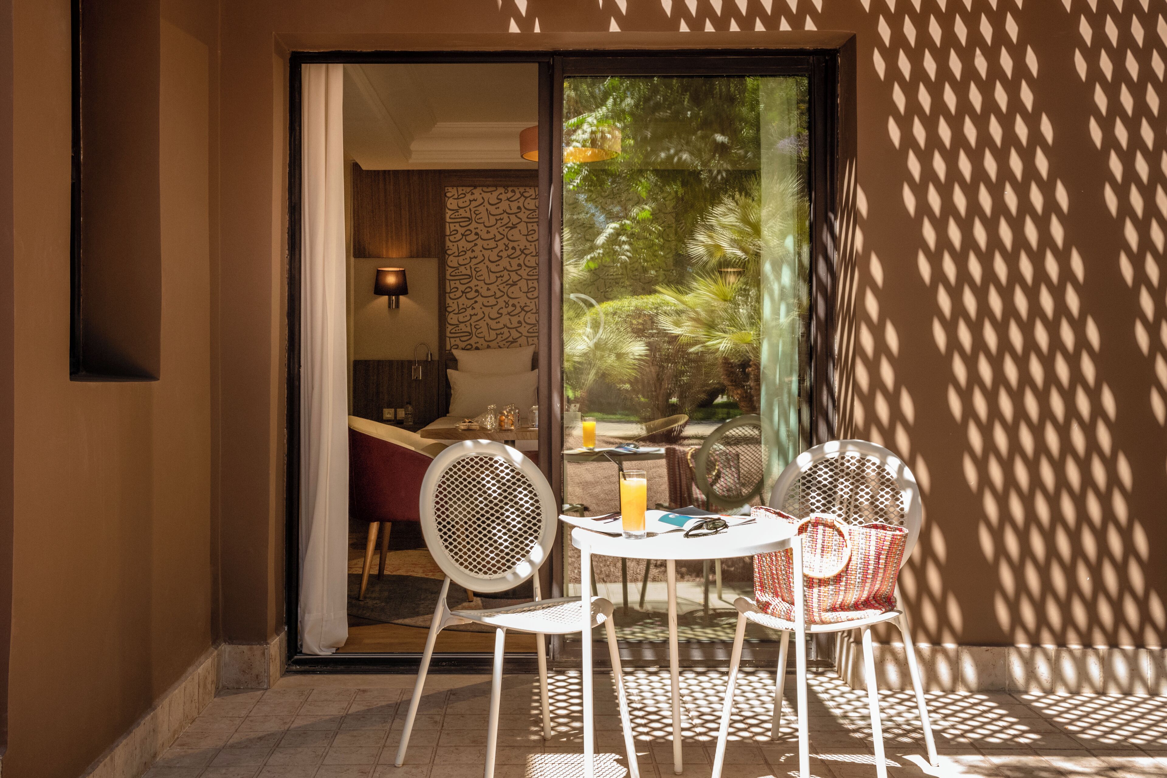 An outdoor seating area with a table and chairs, a glass door leading to a room, and a view of greenery outside.