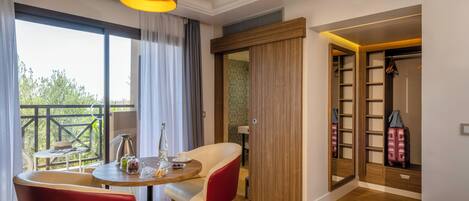 A hotel room with a dining area, a red chair, a wooden wardrobe, and a view of greenery outside.