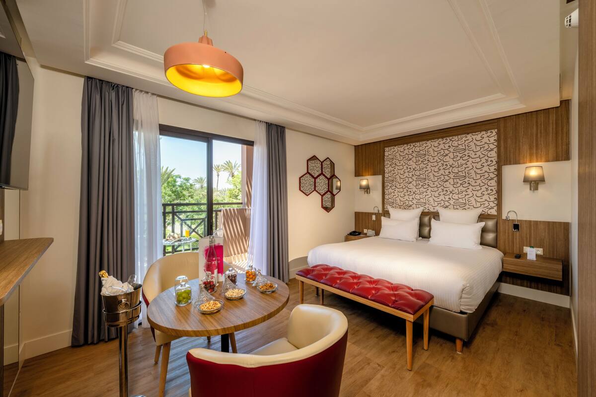 A hotel room with a bed, a dining table with chairs, a red bench, a chandelier, and a view of palm trees.