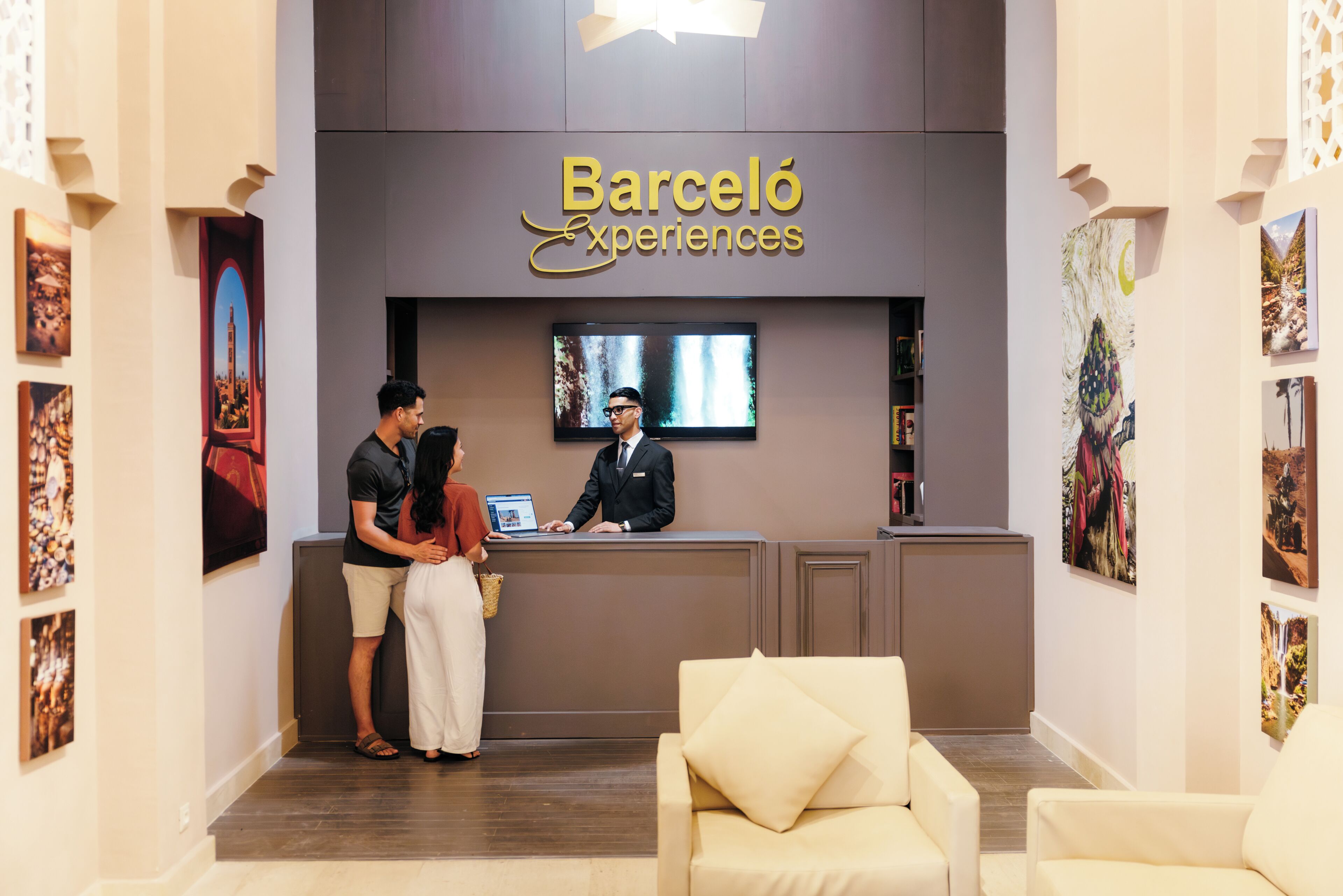 Una reception con una segnaletica che riporta 'Barceló Experiences', un uomo e una donna in piedi accanto al banco, e un divano sullo sfondo.