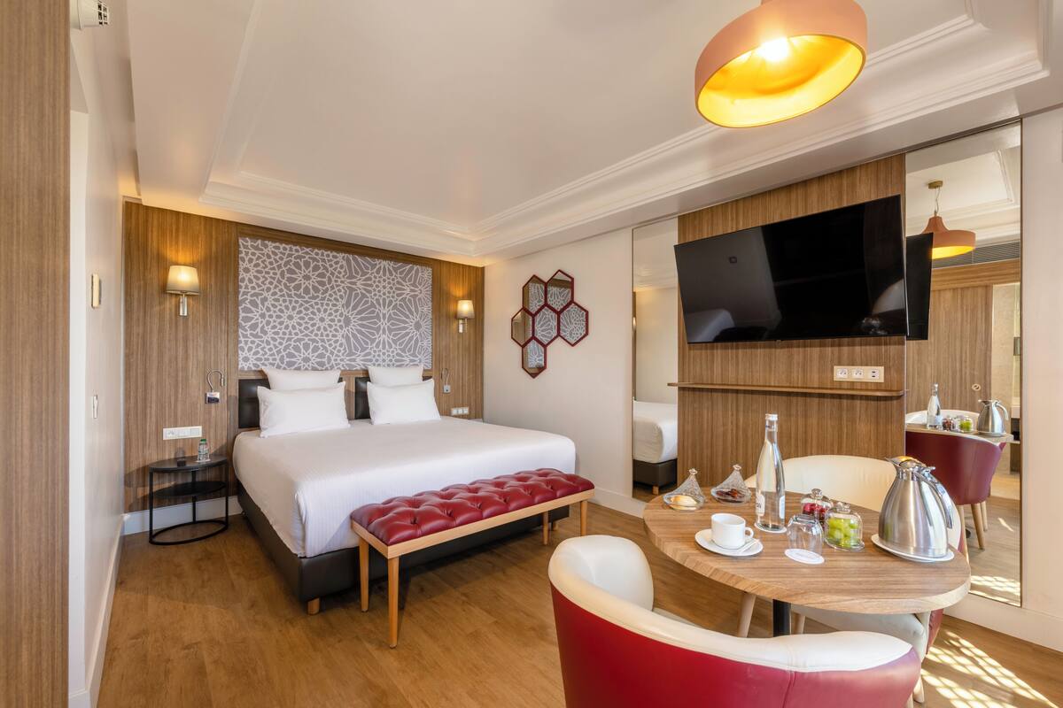 A hotel room with a large bed, a wooden headboard, a flat-screen TV, a dining table with chairs, and a small kitchen area.