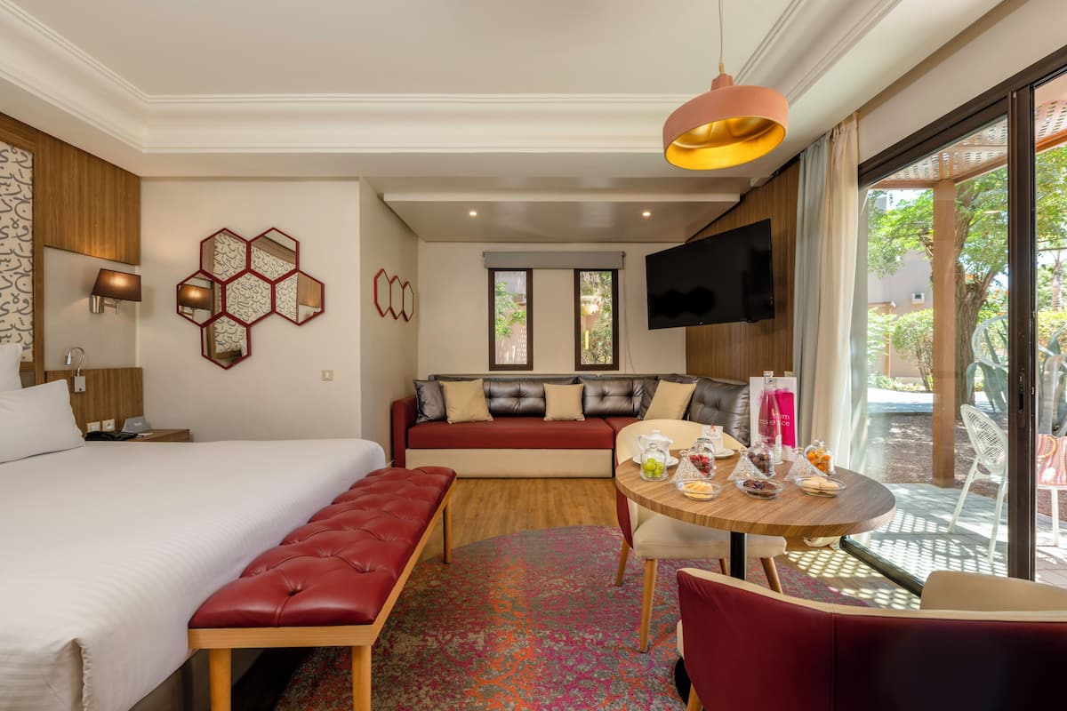 A hotel room with a bed, a sofa, a dining table with chairs, a TV mounted on the wall, and a window with a view of the outdoors.