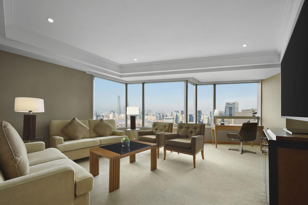 A spacious hotel room with a large window offering a city view, a sofa, a coffee table, and a desk area.