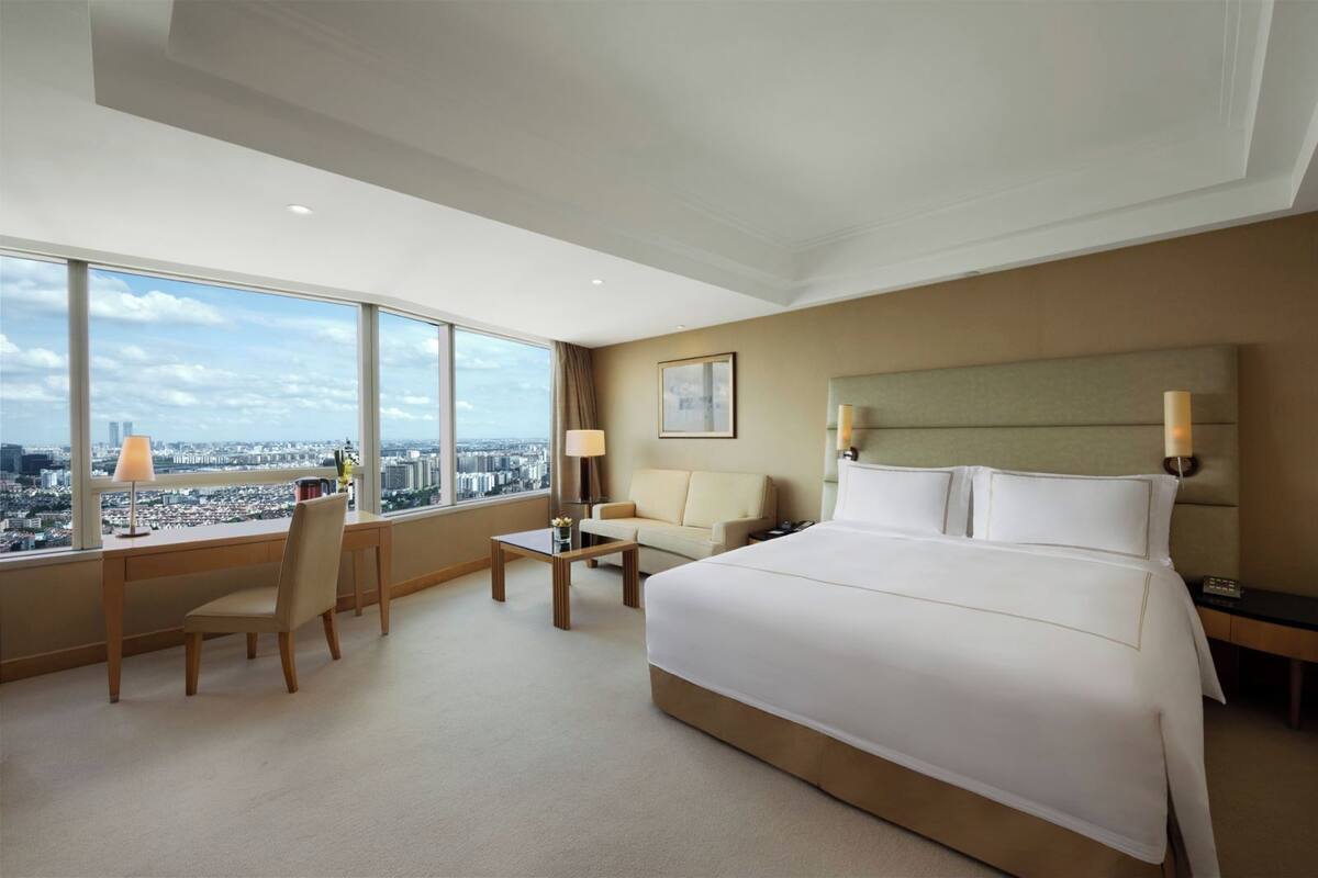 A hotel room with a large bed, a desk, a chair, a small table, and a view of the city through large windows.