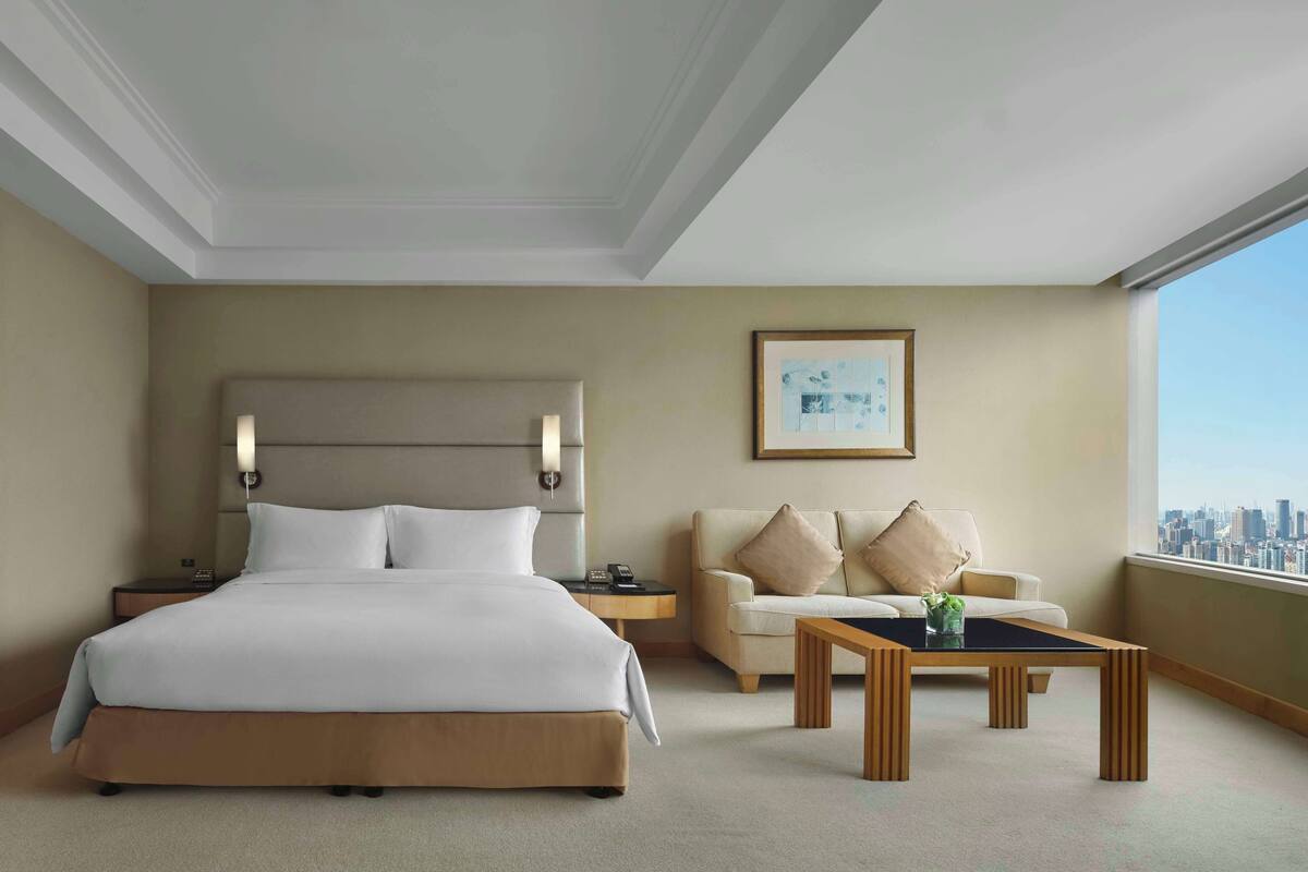 A hotel room with a large bed, a sofa, a coffee table, and a city view.
