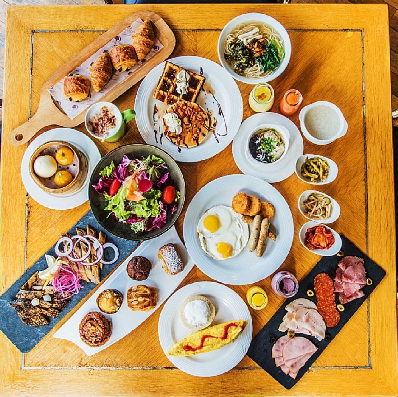 A variety of dishes including a croissant, salad, omelette, and meat platter on a wooden table.