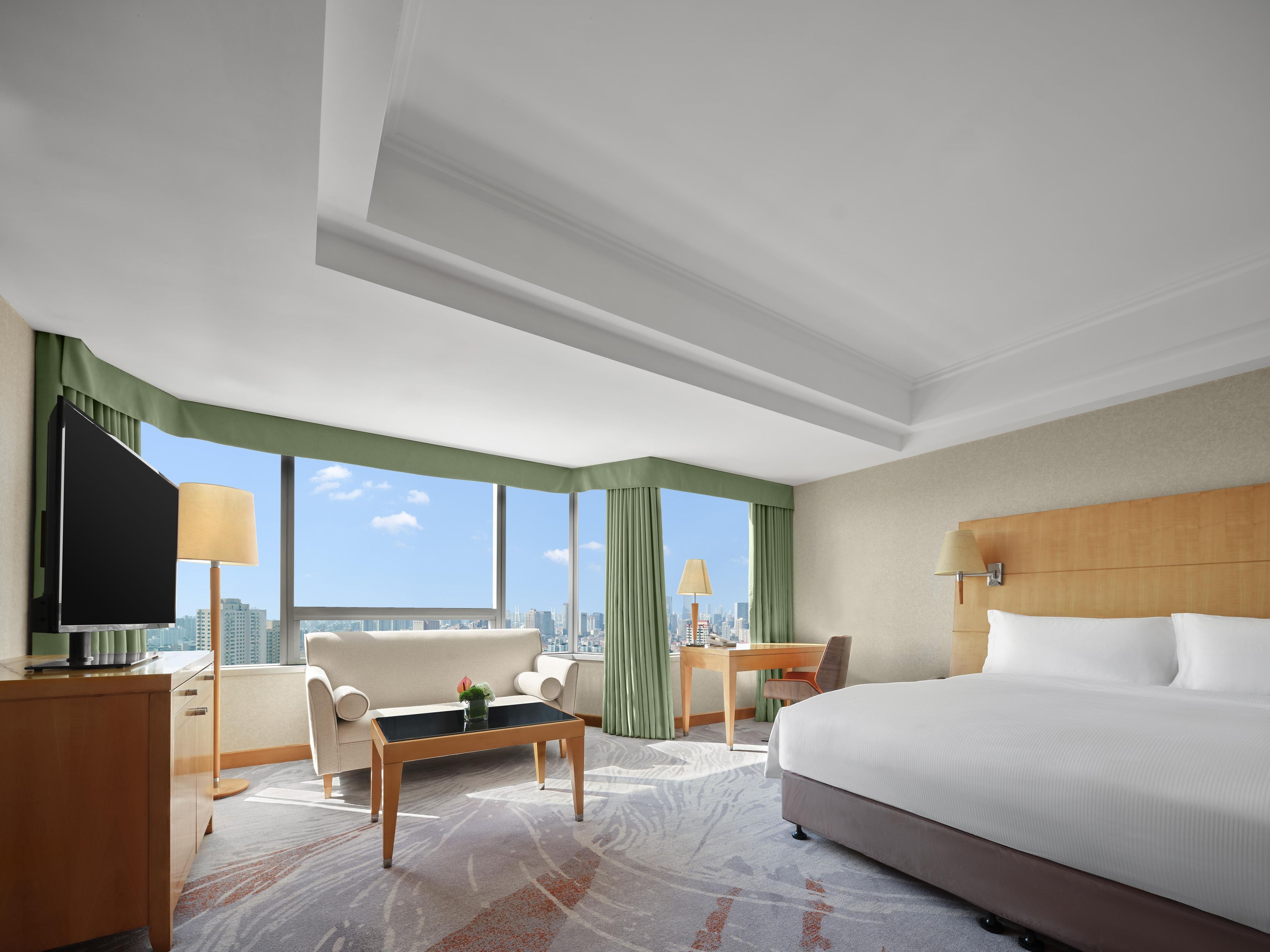 Habitación de hotel con cama, escritorio, silla, sofá, televisor y grandes ventanas con vistas a la ciudad.