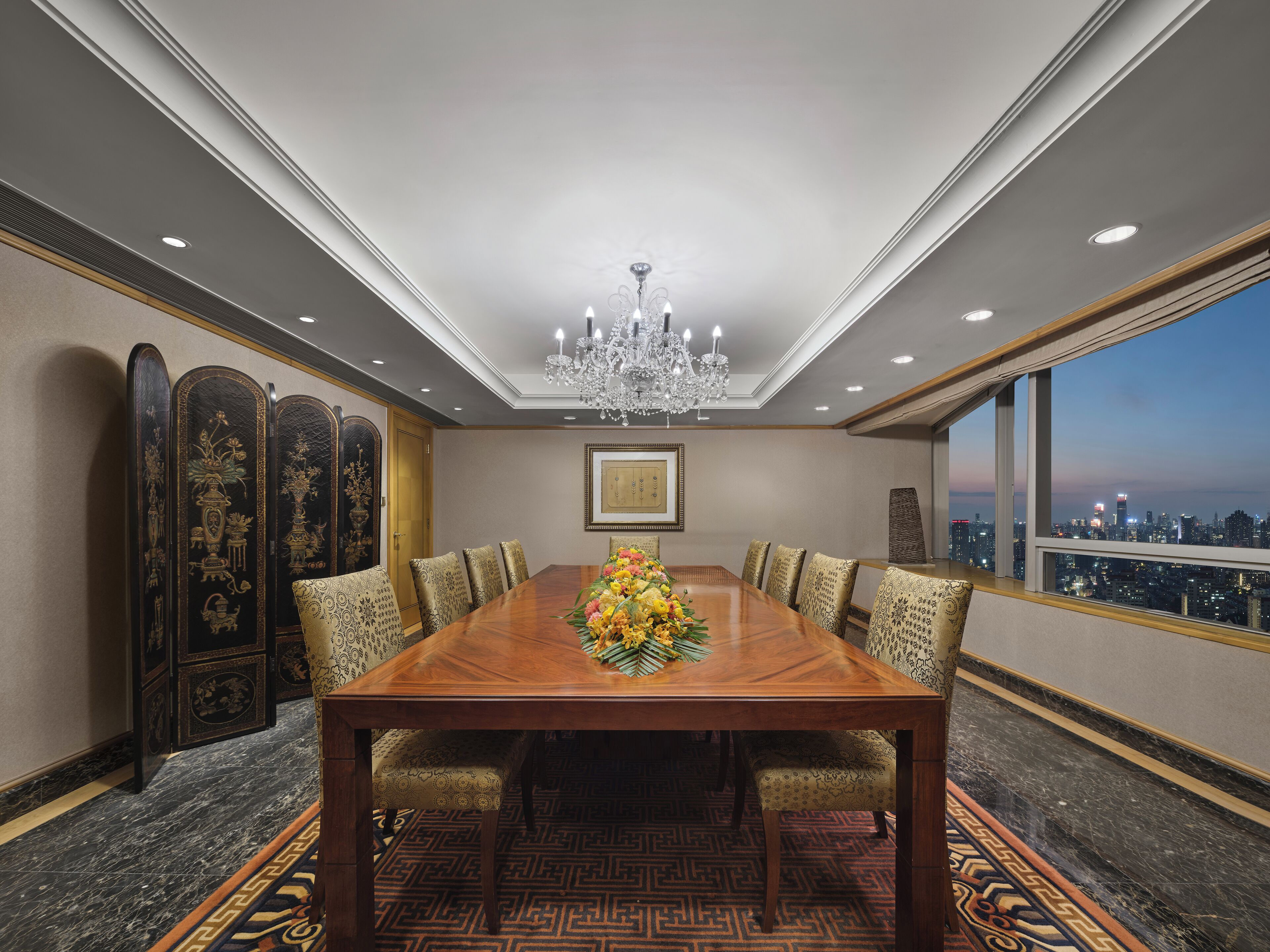 A conference room with a large wooden table, patterned chairs, a chandelier, and a city view through large windows.