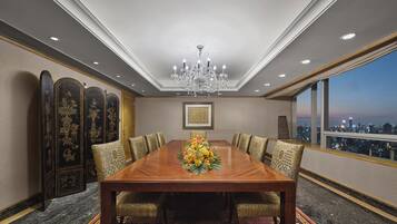 A conference room with a large wooden table, patterned chairs, a chandelier, and a city view through large windows.
