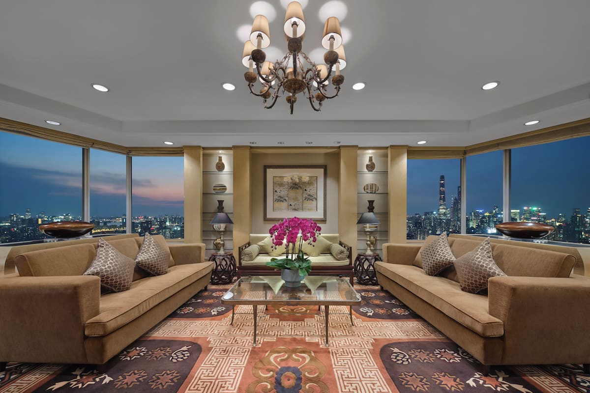A spacious living room with two sofas, a coffee table, and a chandelier. Large windows offer a city view.