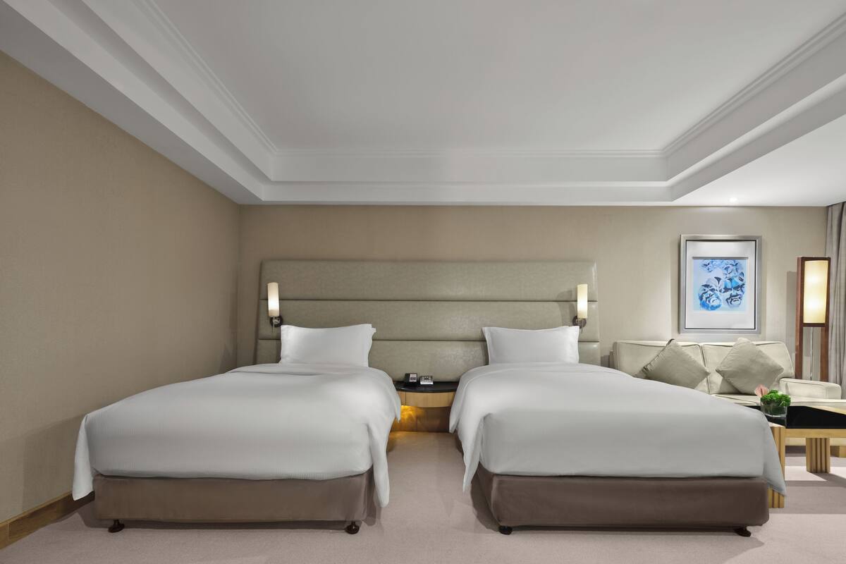 A hotel room with two beds, a headboard, bedside tables, a sofa, and a framed picture on the wall.