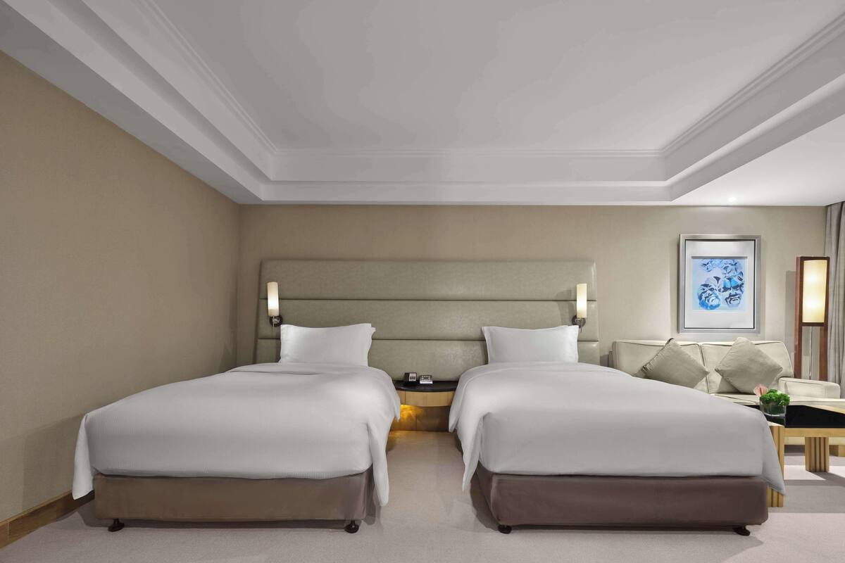 A hotel room with two beds, a large headboard, bedside lamps, a small table with a plant, and a framed picture on the wall.