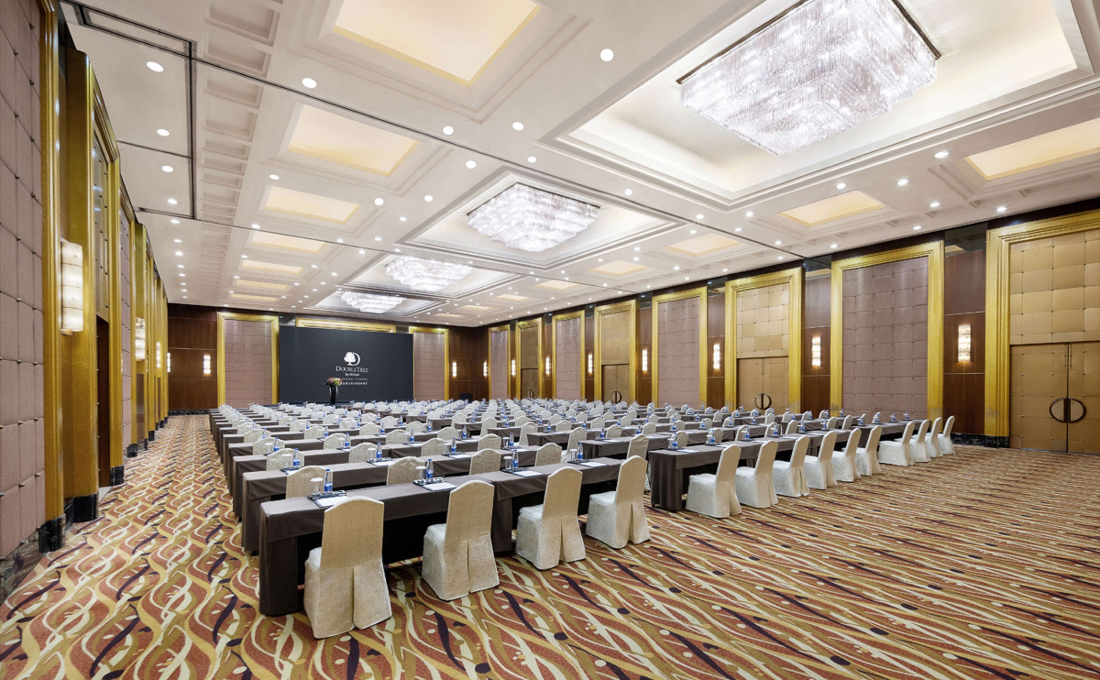 A large conference room with rows of tables and chairs, a stage at the front, and a patterned carpet.