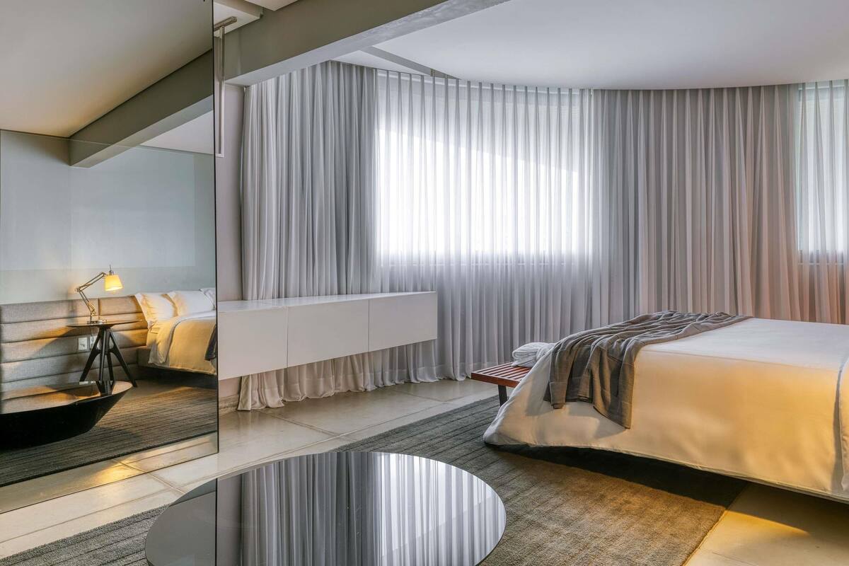 A modern hotel room with a bed, bedside table, lamp, and a large window with curtains.