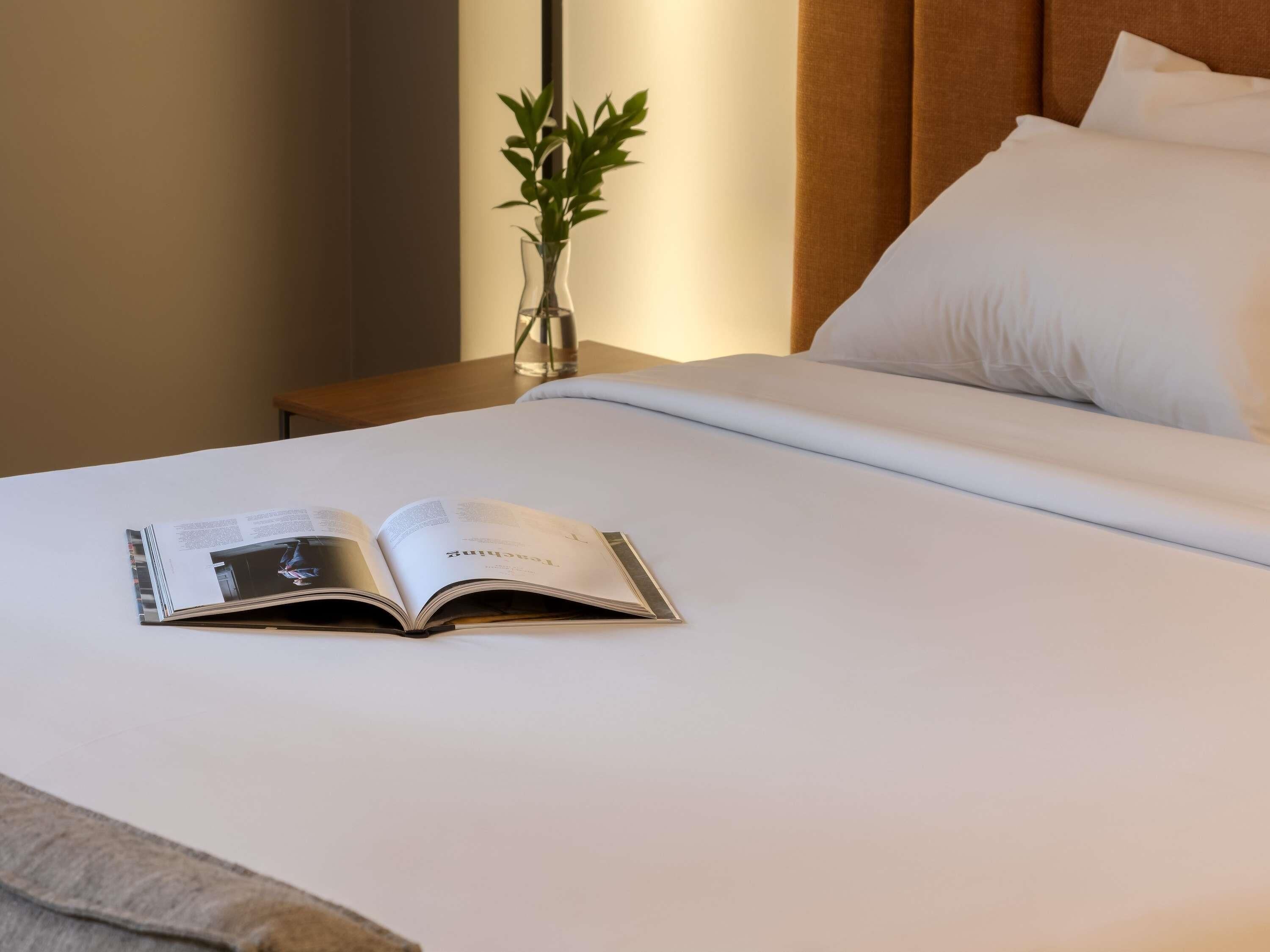 A neatly made bed with white linens, a book open on the bed, and a vase with green plants on a bedside table.