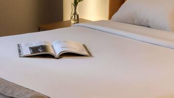 A neatly made bed with white linens, a book open on the bed, and a vase with green plants on a bedside table.