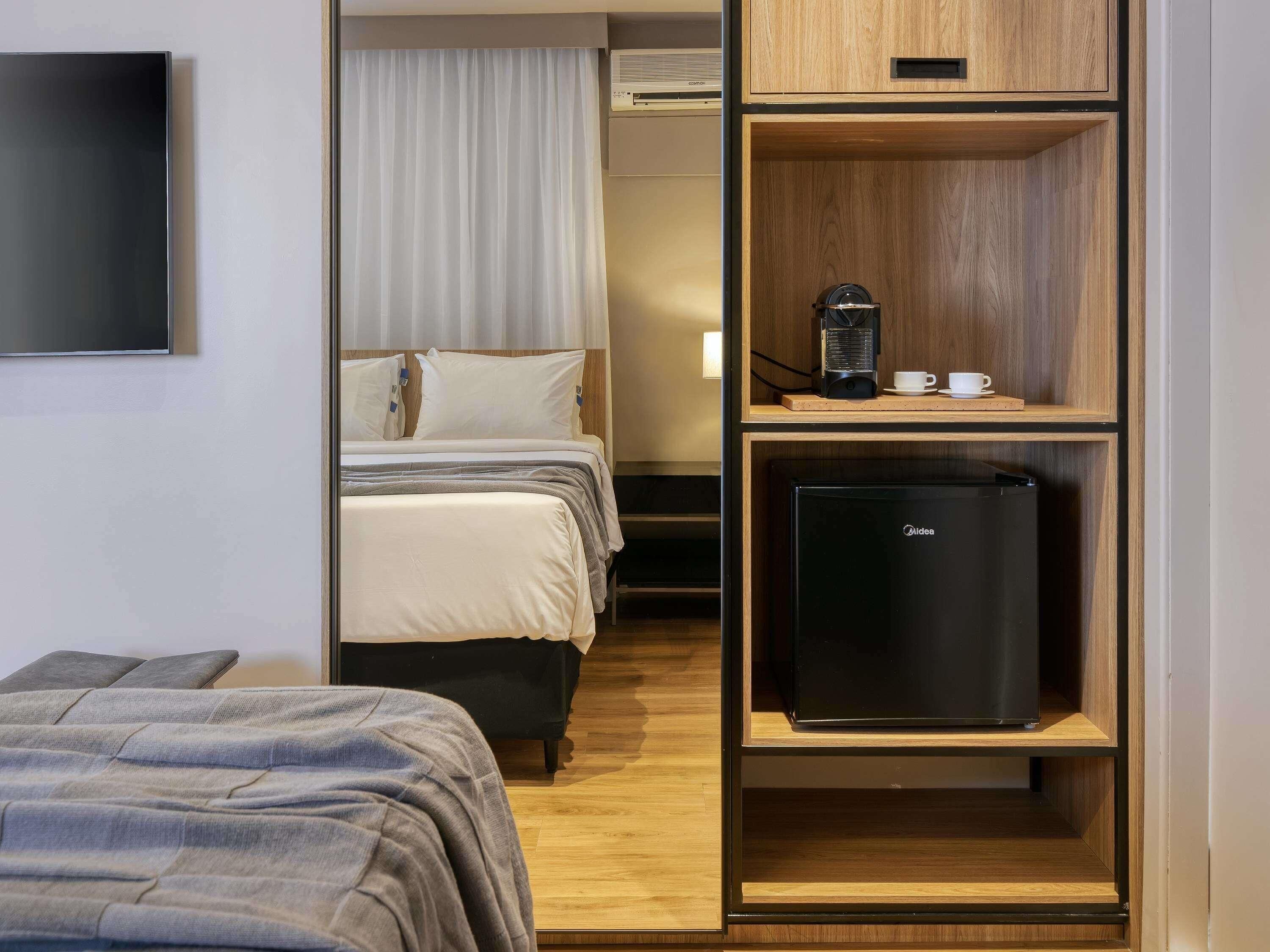 A hotel room with a bed, a desk with a coffee machine, and a refrigerator.