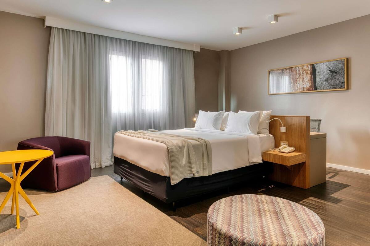 Une chambre d’hôtel moderne avec un grand lit, un bureau, une chaise et un petit pouf.