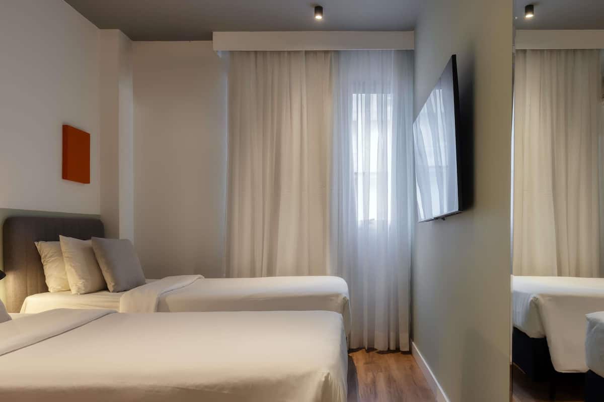 Une chambre d’hôtel avec deux lits, un téléviseur à écran plat fixé au mur et une grande fenêtre avec des rideaux légers.