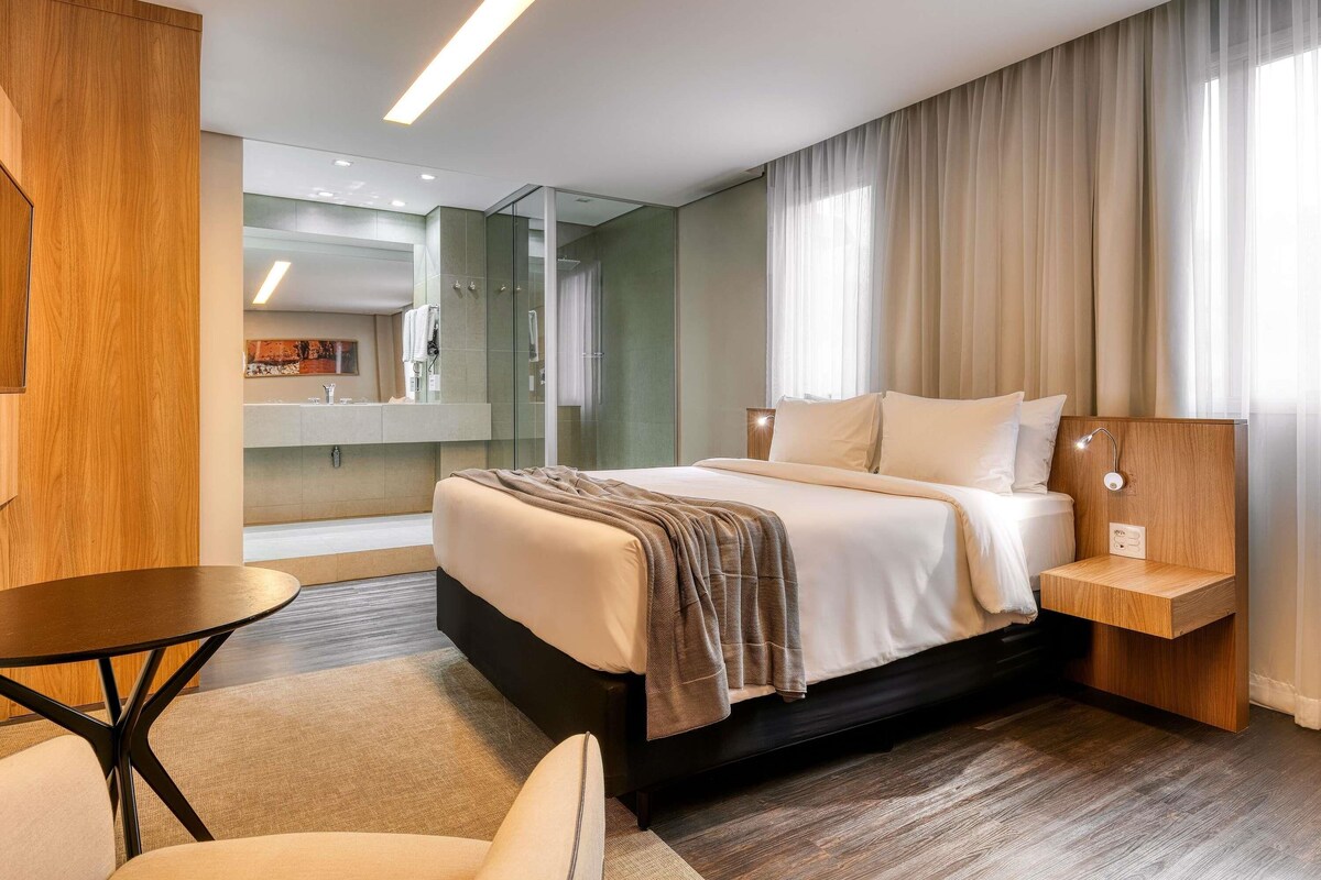 A hotel room with a bed, bedside table, chair, and a bathroom visible through a glass partition.