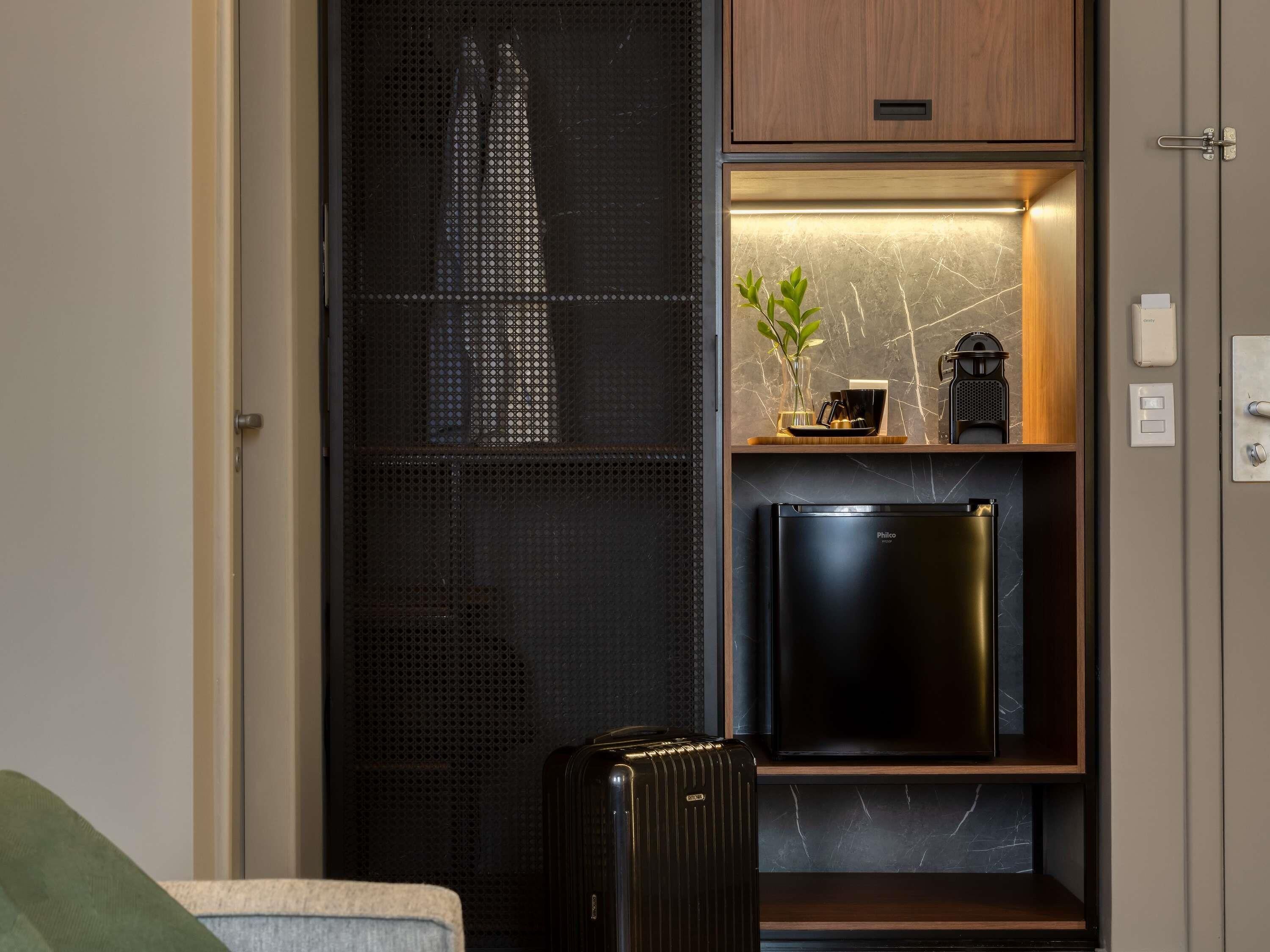 A compact kitchenette with a black refrigerator, a small shelf with a plant and a coffee maker, and a suitcase on the floor.
