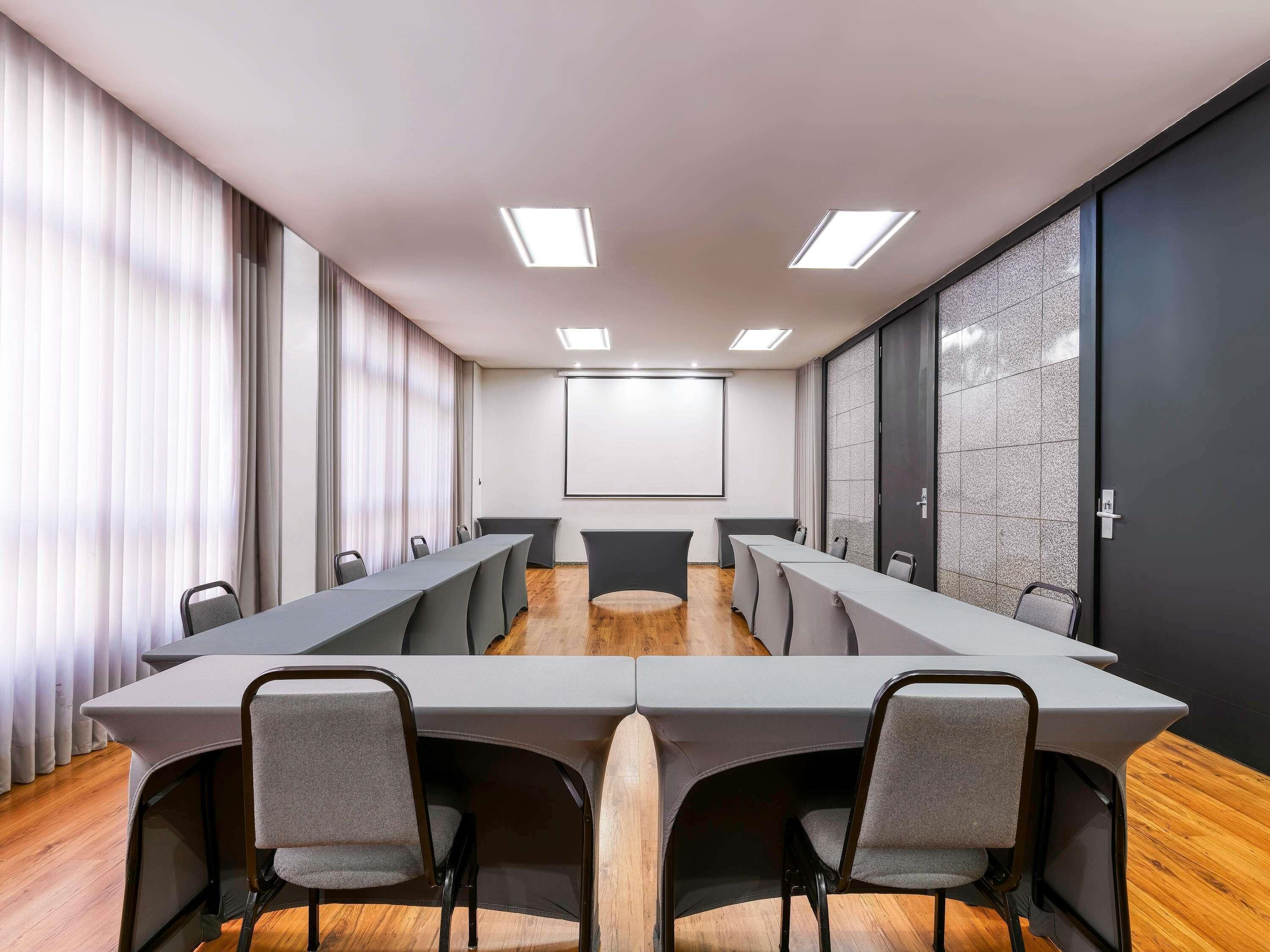 A conference room with a long table, chairs, a projector screen, and a large window with curtains.