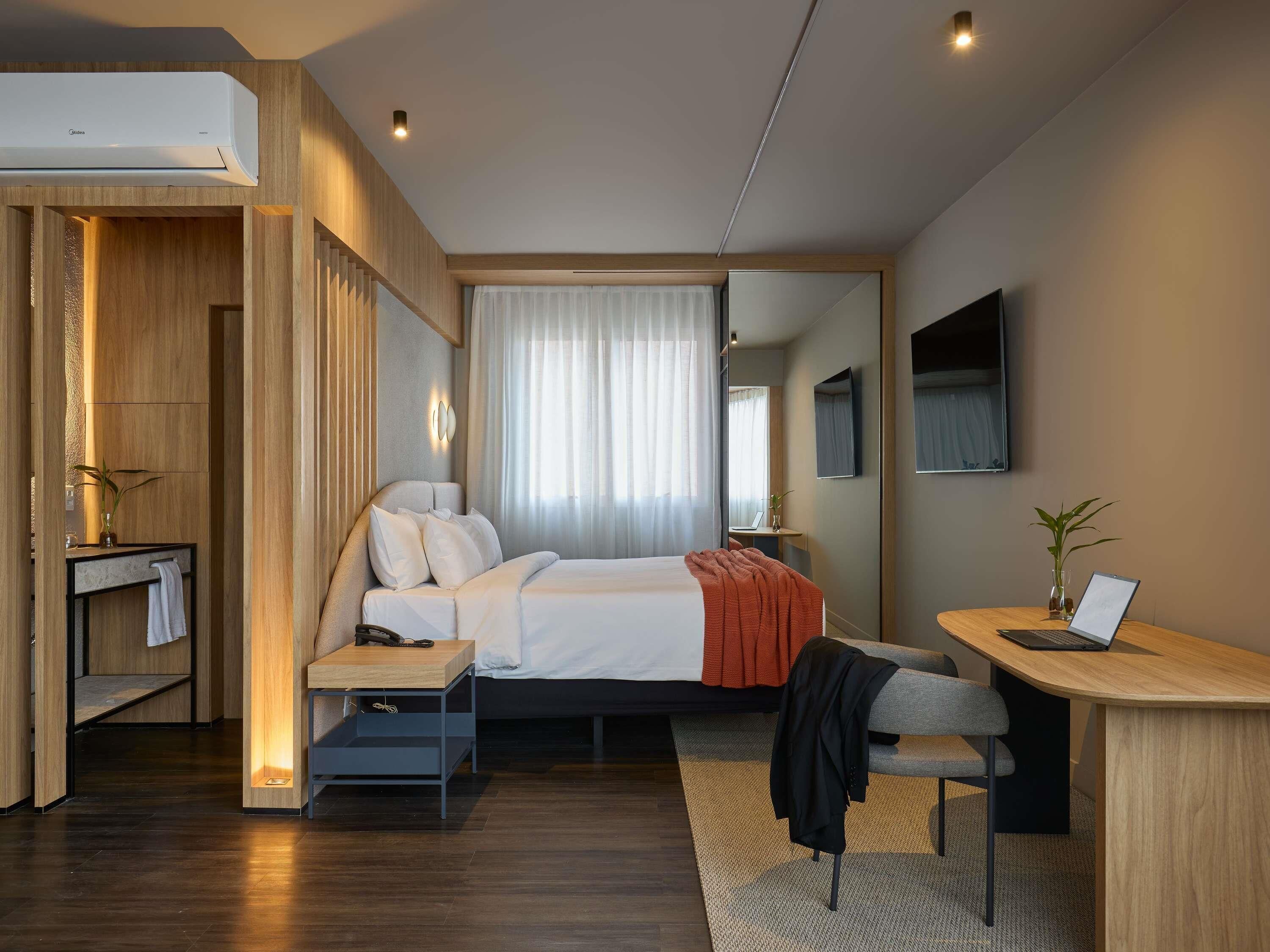 Quarto de hotel moderno com cama, mesa de cabeceira, escrivaninha e um espelho grande.