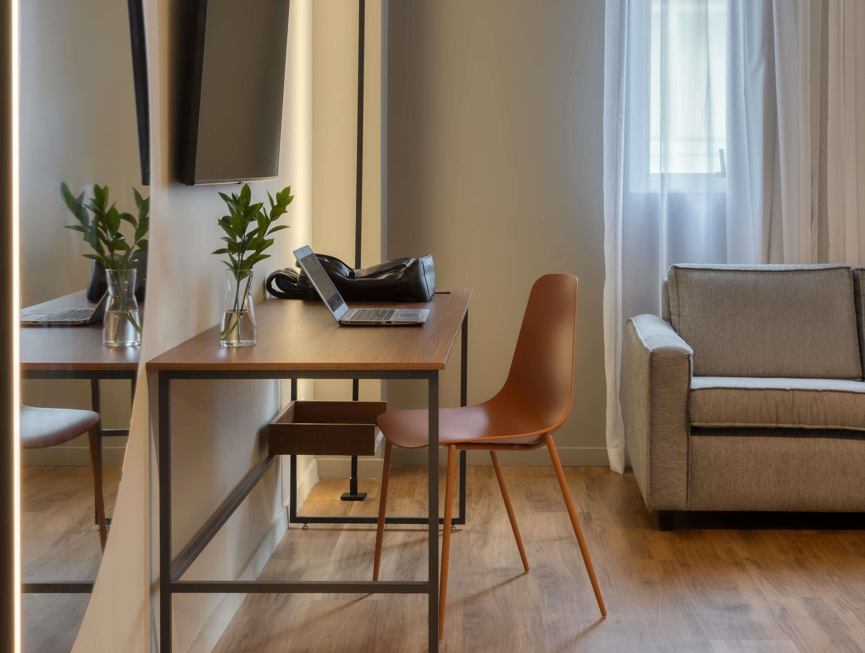 A modern living room with a wooden desk, a laptop, a chair, a sofa, and two potted plants.