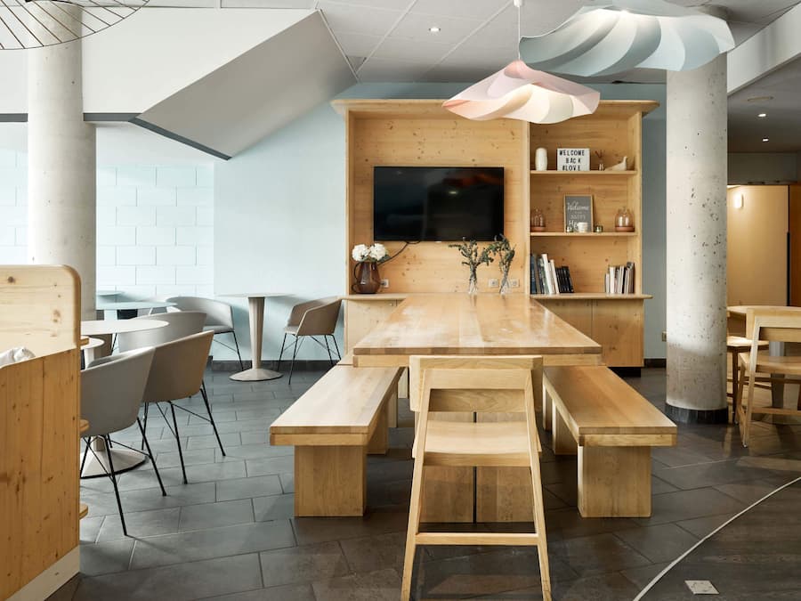 A modern, open-plan lounge area with a wooden table and benches, a flat-screen TV mounted on a wooden wall, and a bookshelf with various items.