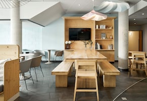 Un salón moderno de planta abierta con una mesa de madera y bancos, un televisor de pantalla plana montado en una pared de madera, y una estantería con diversos objetos.