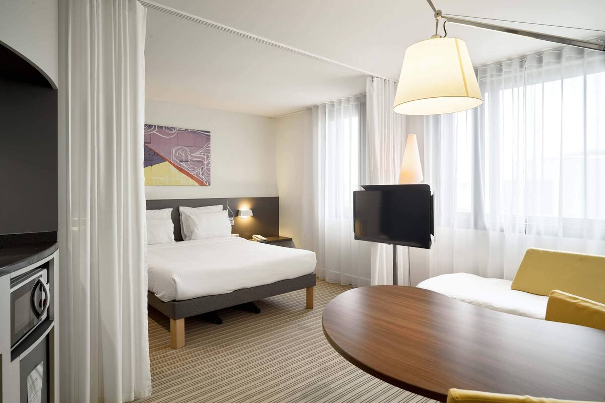 A hotel room with a bed, bedside table, lamp, TV, and a dining area with a table and chairs.
