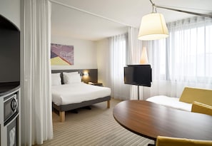 A hotel room with a bed, bedside table, lamp, TV, and a round table with yellow chairs.