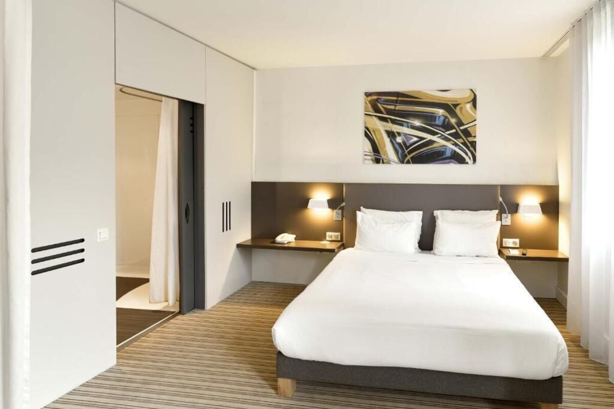 A hotel room with a bed, bedside tables, a painting, and a wardrobe.