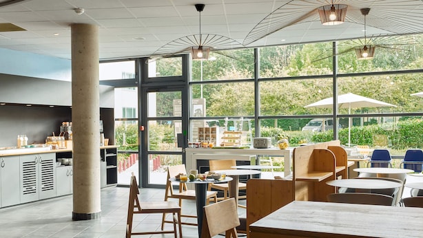 A modern cafe with large windows, wooden furniture, and a visible kitchen area. - B&B HOTEL Paris Grand Roissy CDG Aéroport (Roissy-en-France)
