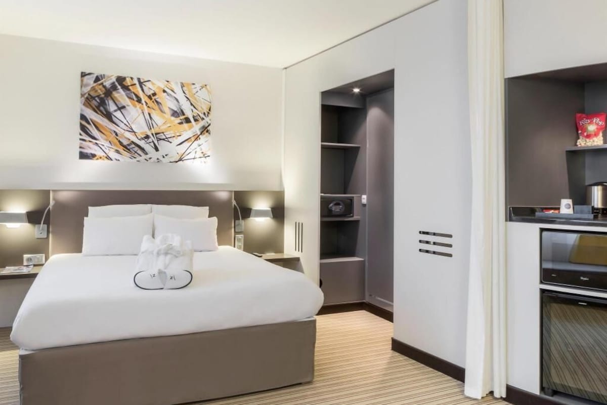 A hotel room with a bed, bedside tables, a wall-mounted artwork, a built-in wardrobe, and a kitchenette.