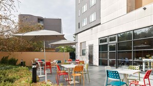 An outdoor patio with multiple tables and colorful chairs, surrounded by a garden and a modern building with large windows. - B&B HOTEL Paris Grand Roissy CDG Aéroport (Roissy-en-France)