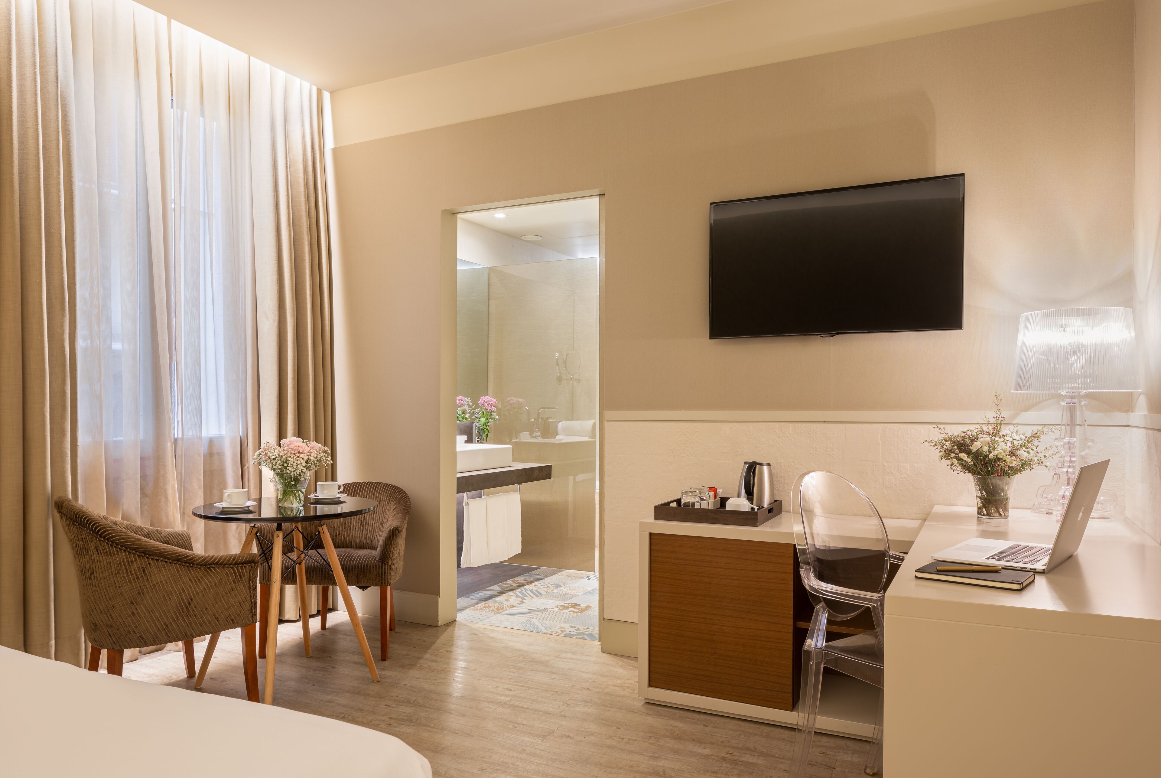 Habitación de hotel con cama, escritorio con laptop, silla, televisor y baño visible a través de una puerta abierta.