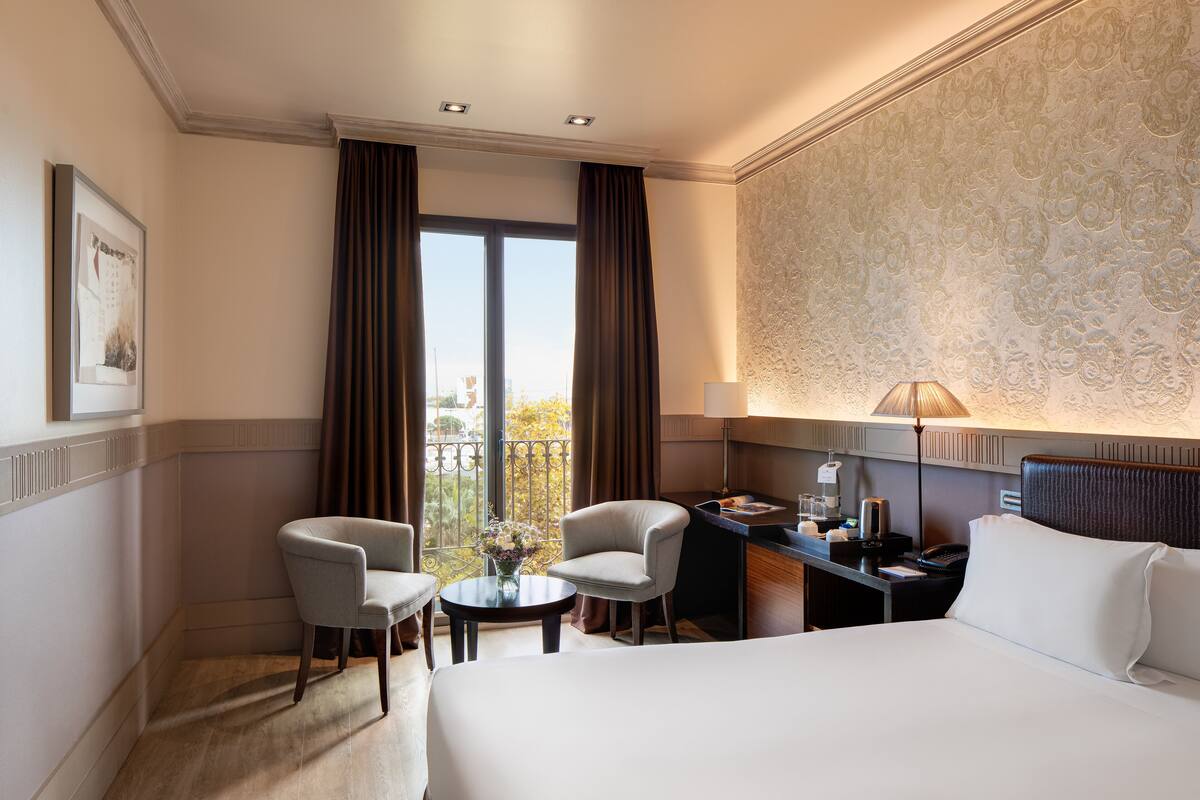 Ein Hotelzimmer mit einem Bett, zwei Sesseln, einem Schreibtisch mit Lampe und Blick auf die Stadt durch große Fenster.