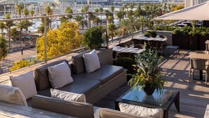 Una terraza en la azotea con mobiliario exterior, vistas al puerto deportivo y un paisaje urbano de fondo.