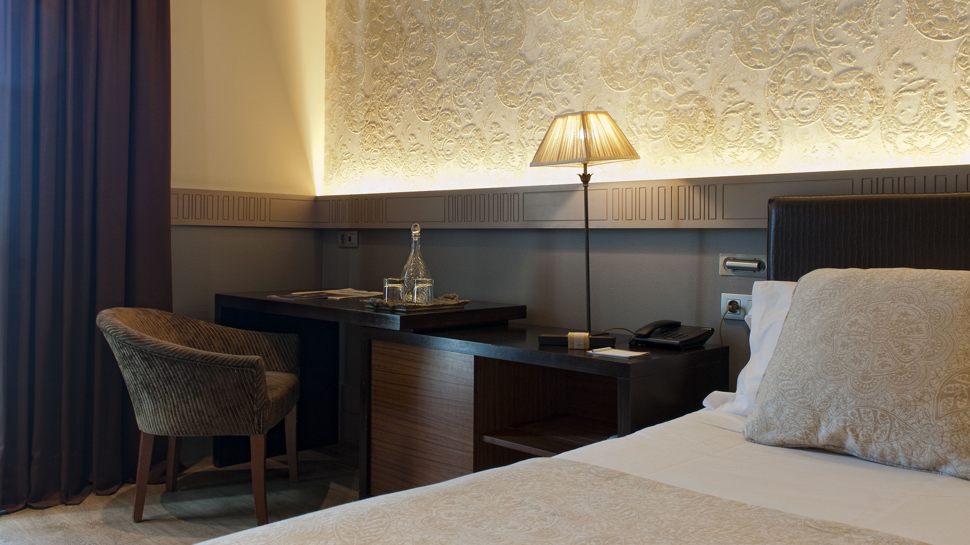 A hotel room with a bed, a desk with a lamp, a telephone, and a chair.