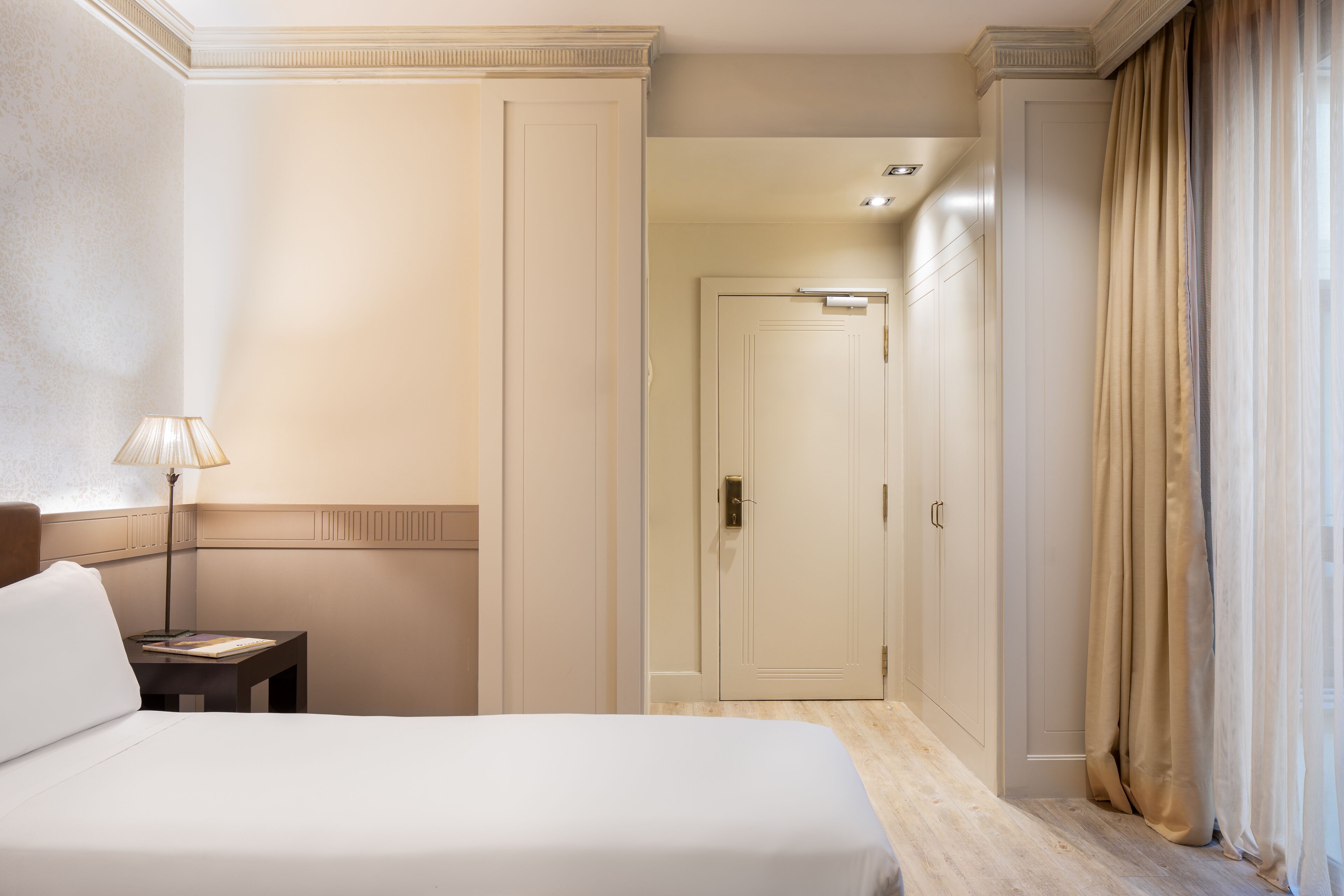 Una habitación de hotel con cama, mesita de noche, lámpara, puerta y ventana con cortinas.