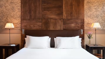 A neatly made bed with white linens and two pillows, flanked by two bedside tables with lamps and a vase of flowers.