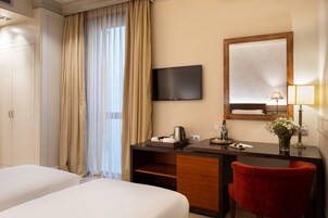 A hotel room with two beds, a desk with a flat-screen TV, a red chair, a mirror, and a vase with flowers.