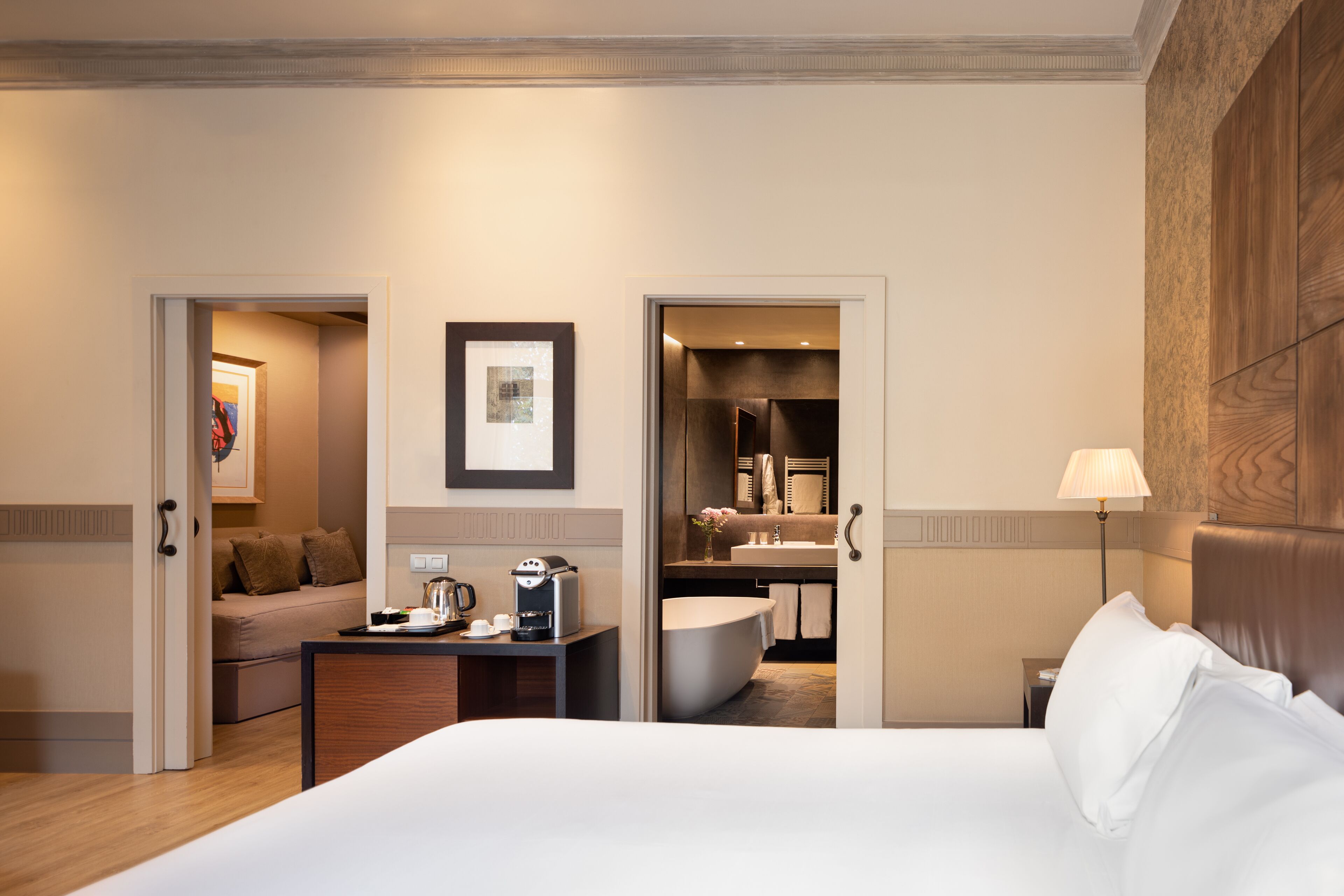 A hotel room with a bed, a desk with a coffee maker, a sofa, a bathroom with a bathtub, and a framed picture on the wall.
