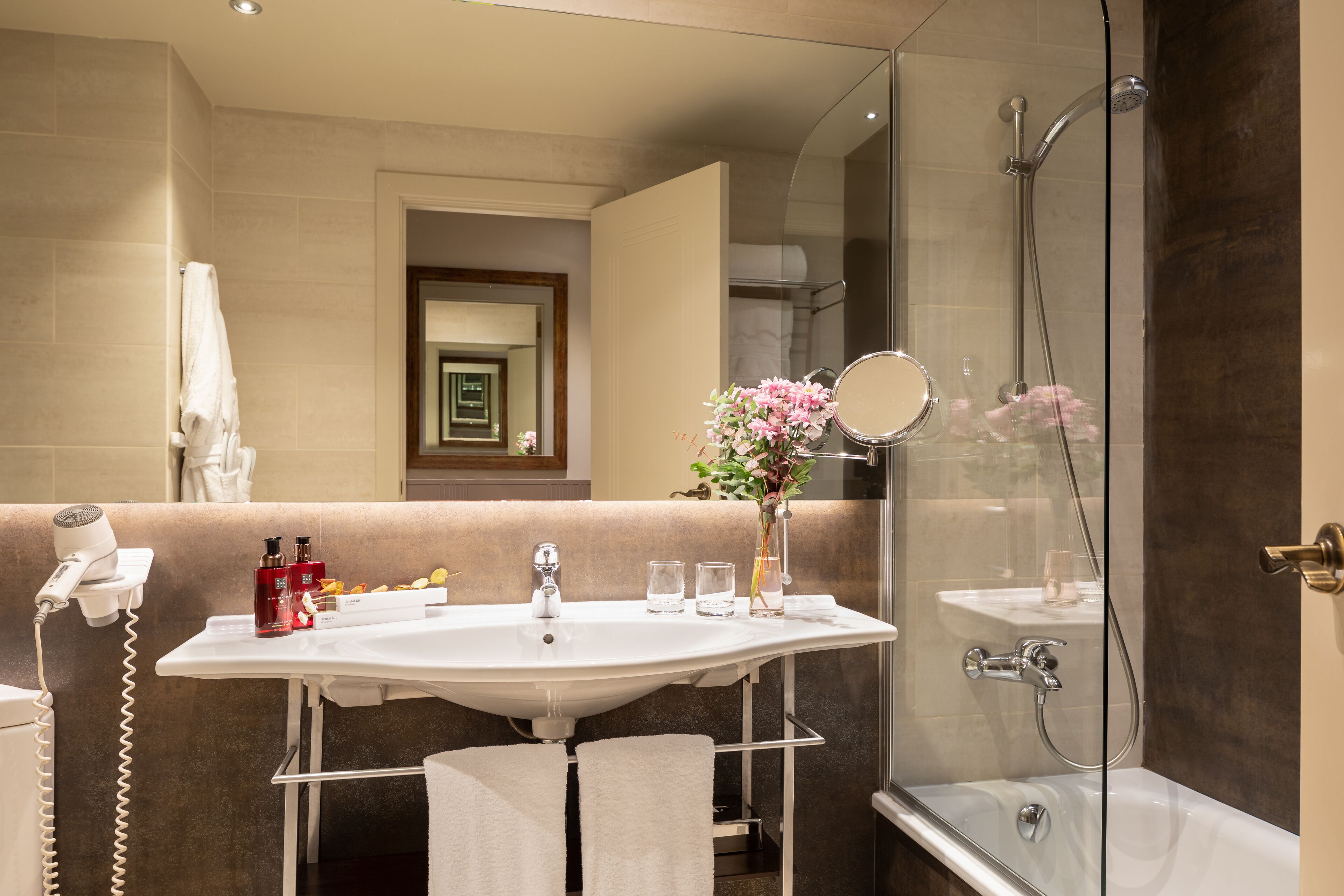 Un baño moderno con ducha envuelta en vidrio, lavamanos blanco con espejo y zona de tocador con artículos de aseo.