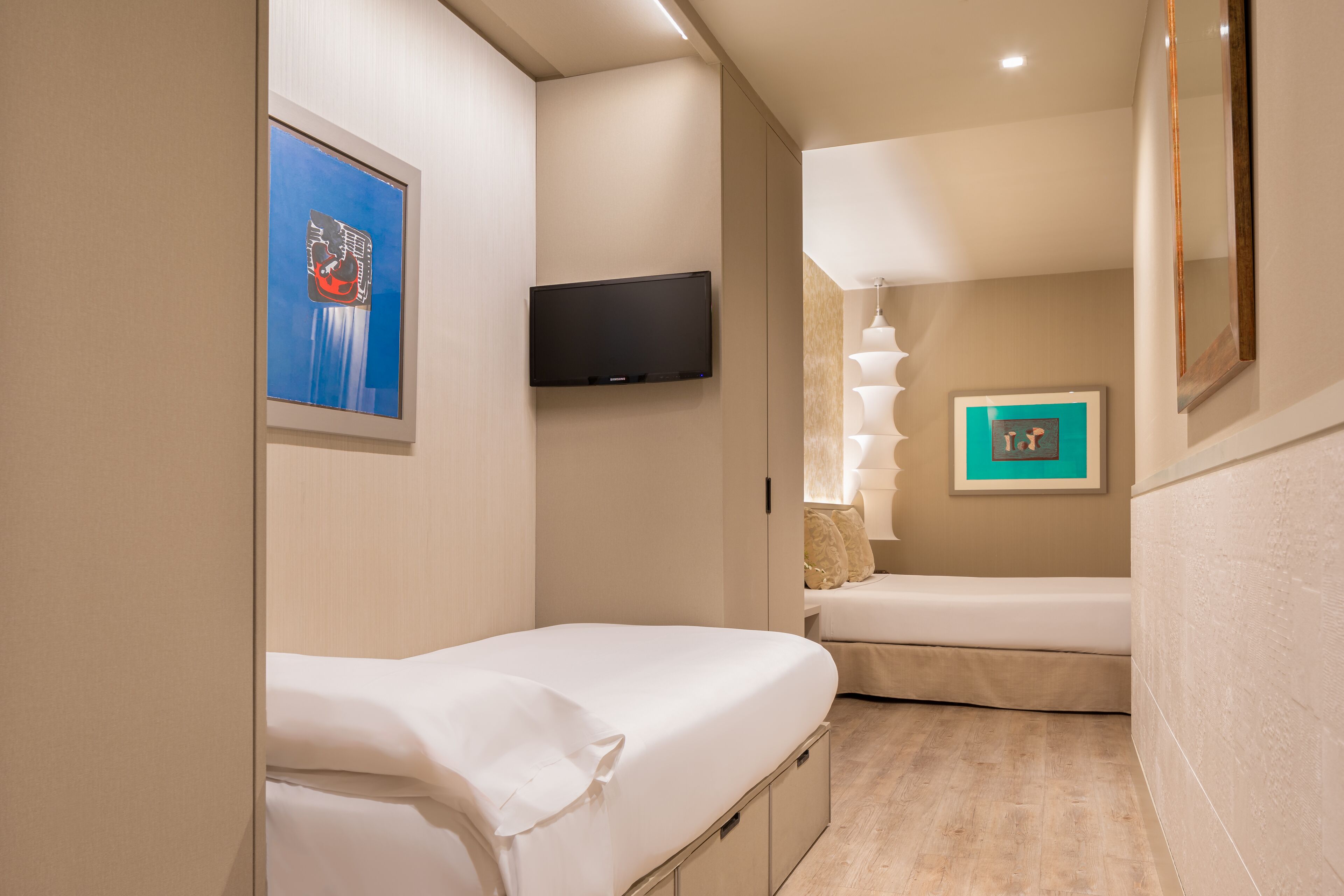 A compact hotel room with a single bed, a television mounted on the wall, and a small seating area with a lamp.