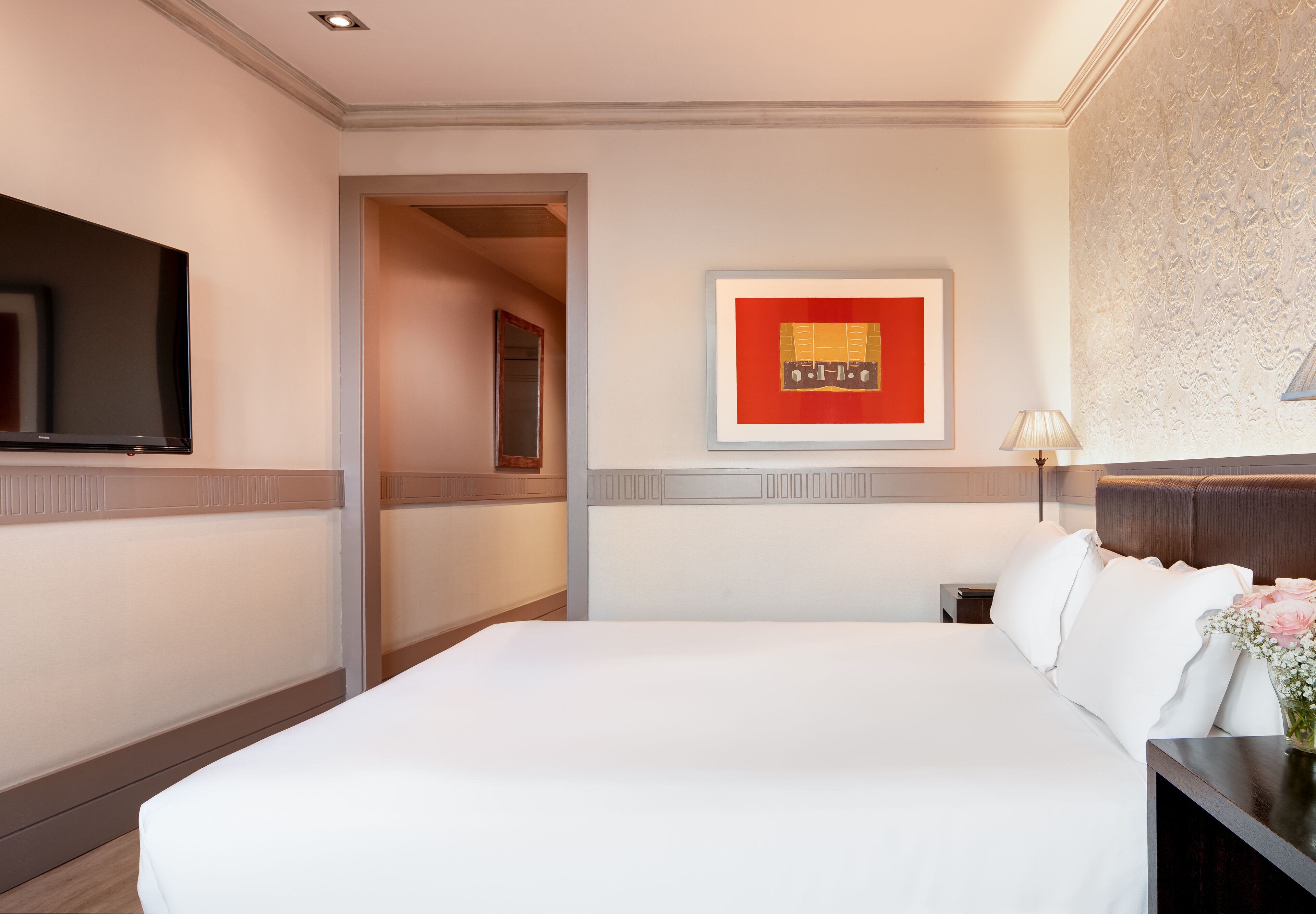 Une chambre d’hôtel avec un grand lit, un téléviseur à écran plat fixé au mur, une œuvre d’art encadrée au-dessus du lit et une table de chevet avec une lampe.